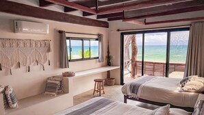 A bedroom with two beds, a large window with a view of the ocean, a macrame wall hanging, and a ceiling fan. - Amansala (Tulum)