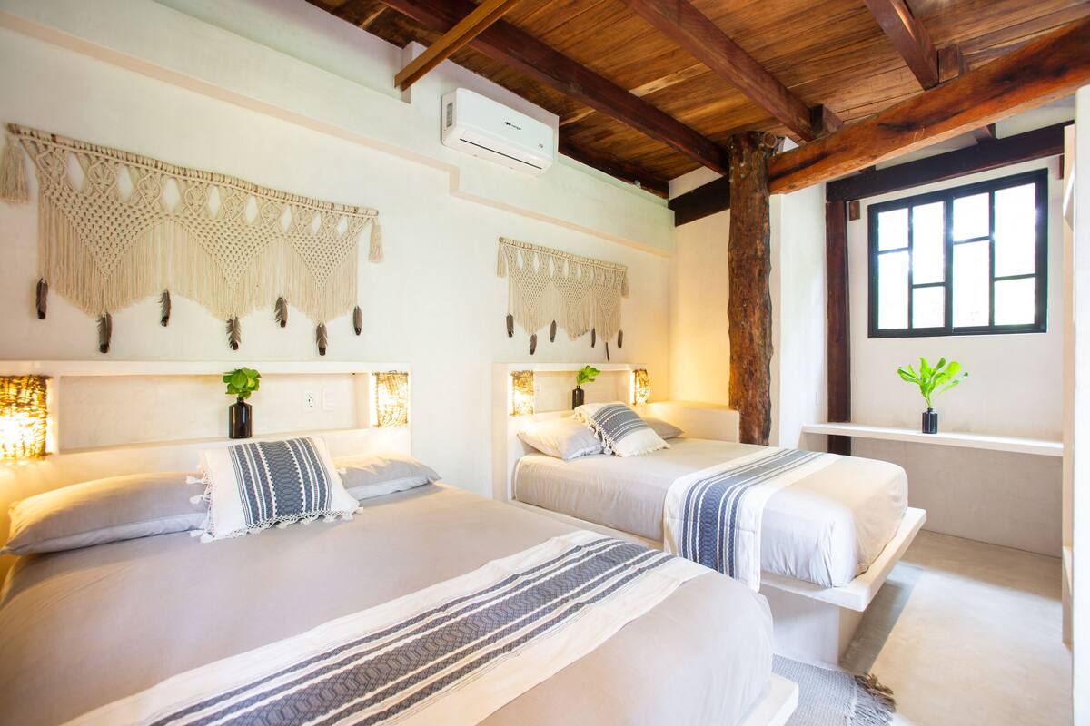 A hotel room with two beds, macramé wall hangings, a ceiling fan, and a window with a view.