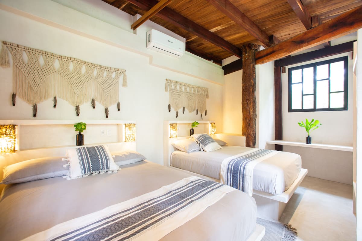 A hotel room with two beds, macramé wall hangings, a ceiling fan, and a window with a view.