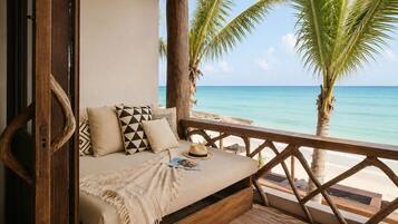 Terraza de madera con sofá cama, almohadas y un sombrero, con vistas a una playa con palmeras y agua azul cristalina.