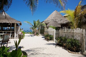 Una propiedad frente a la playa con cabañas de techo de paja, un sendero arenoso y plantas tropicales.