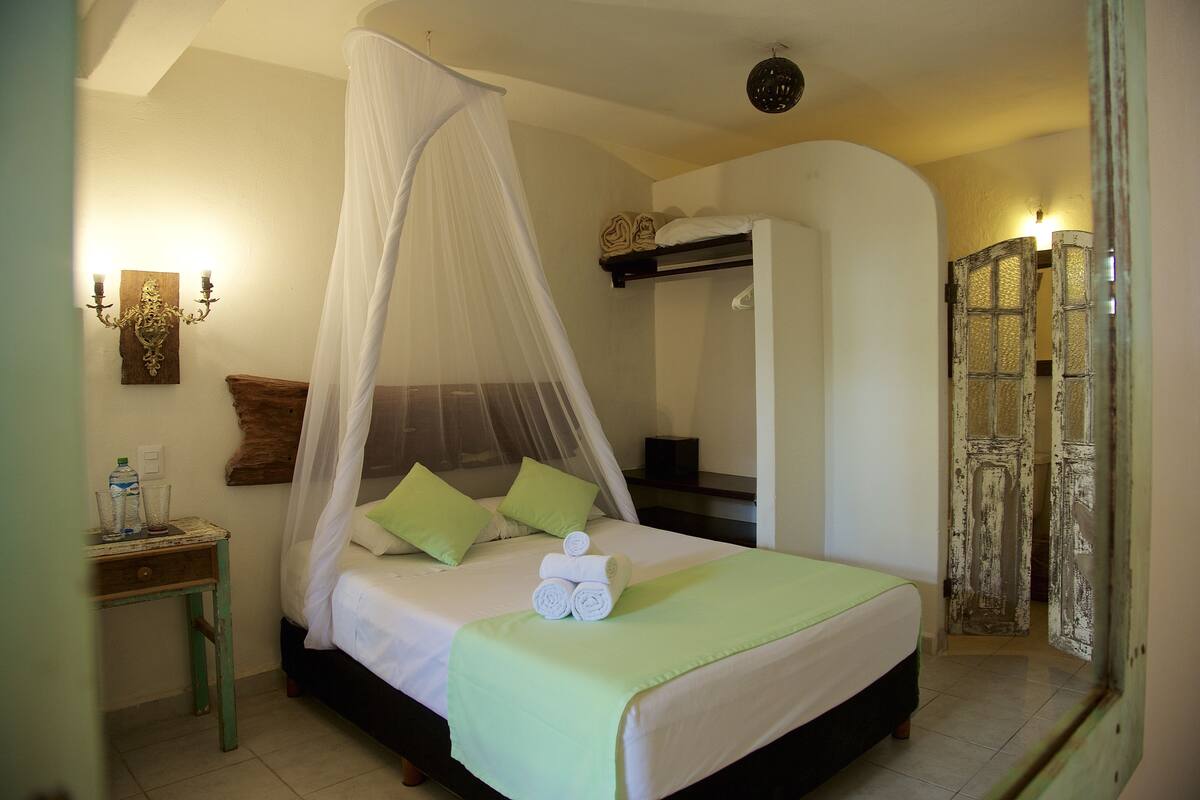 A neatly made bed with a canopy, a bedside table with a water bottle and glasses, and a rustic door in the background.