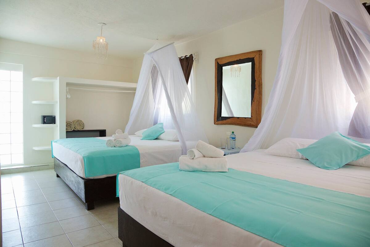 Two beds with white and teal bedding, a wooden headboard, a bedside table with a bottle, and a white ceiling light.