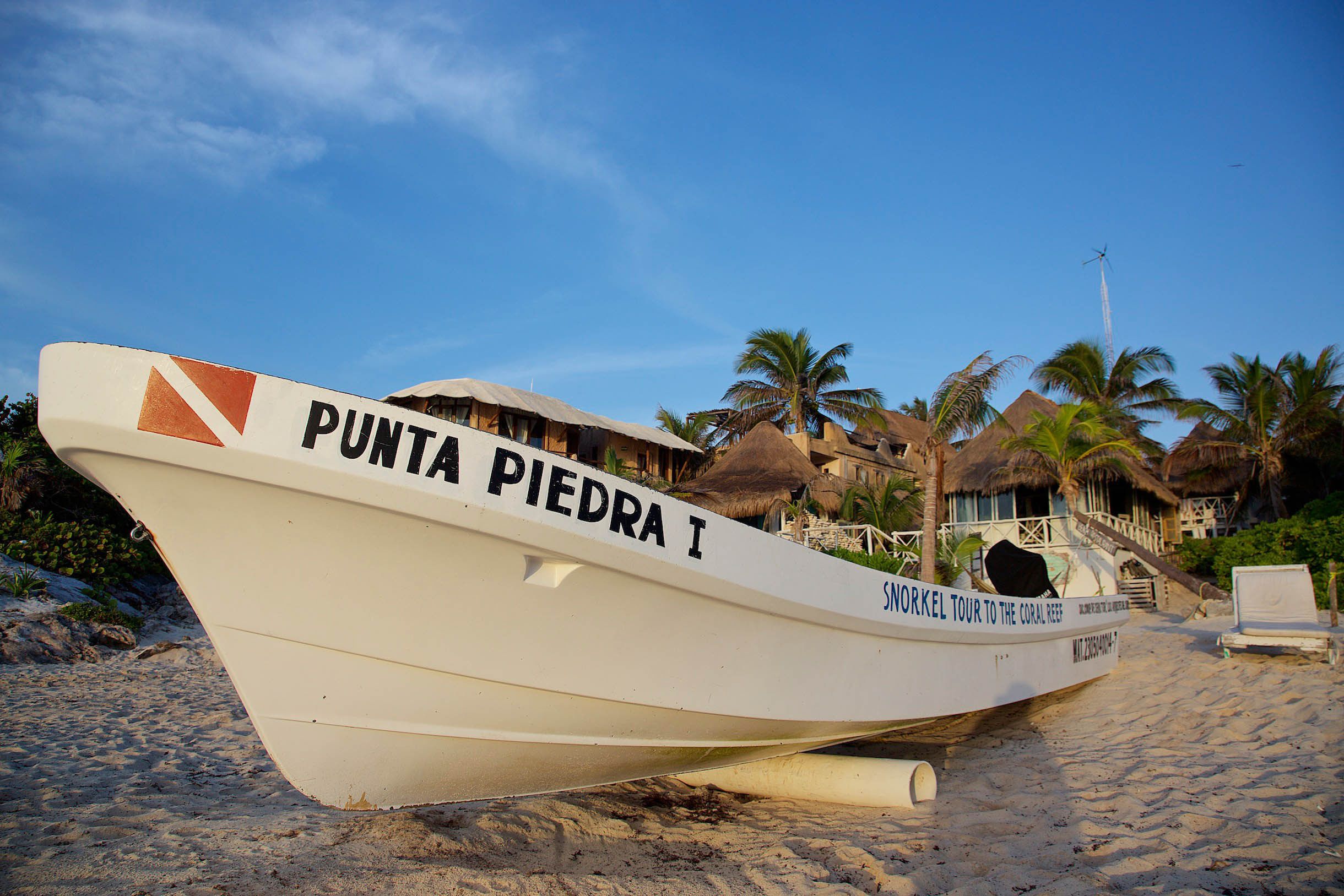 Un bote llamado Punta Piedra I sobre una playa arenosa, con estructuras de techo de paja y palmeras al fondo.
