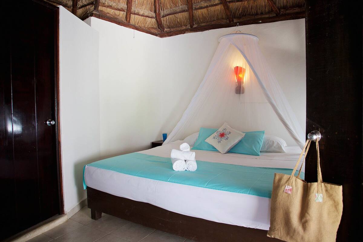 A neatly made bed with a white and teal bedspread, a rolled towel, and a single pillow. A mosquito net is draped over the bed, and a bag hangs on the wall.