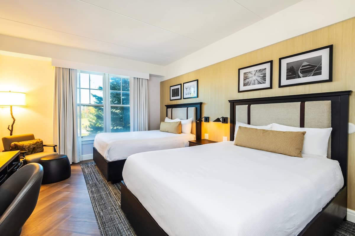 A hotel room with two beds, a desk, a chair, a lamp, and a window with curtains.