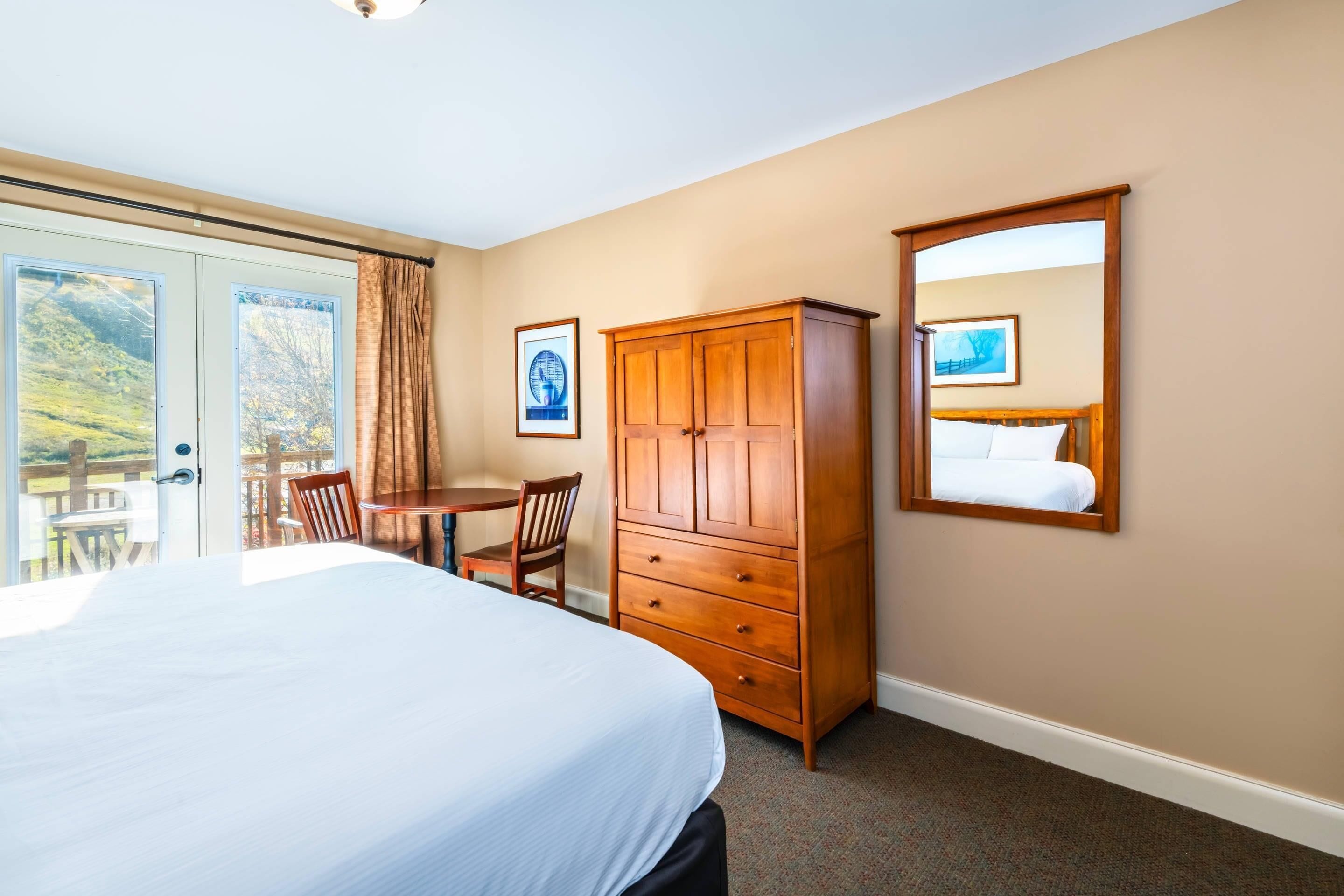 Una habitación de hotel con una cama, un armario de madera, una mesa de comedor con sillas y un ventanal grande.