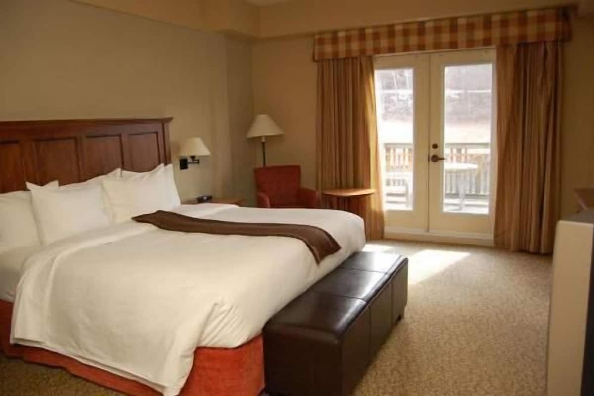 A hotel room with a large bed, a bench, a chair, a lamp, and a view of the outdoors through a door.