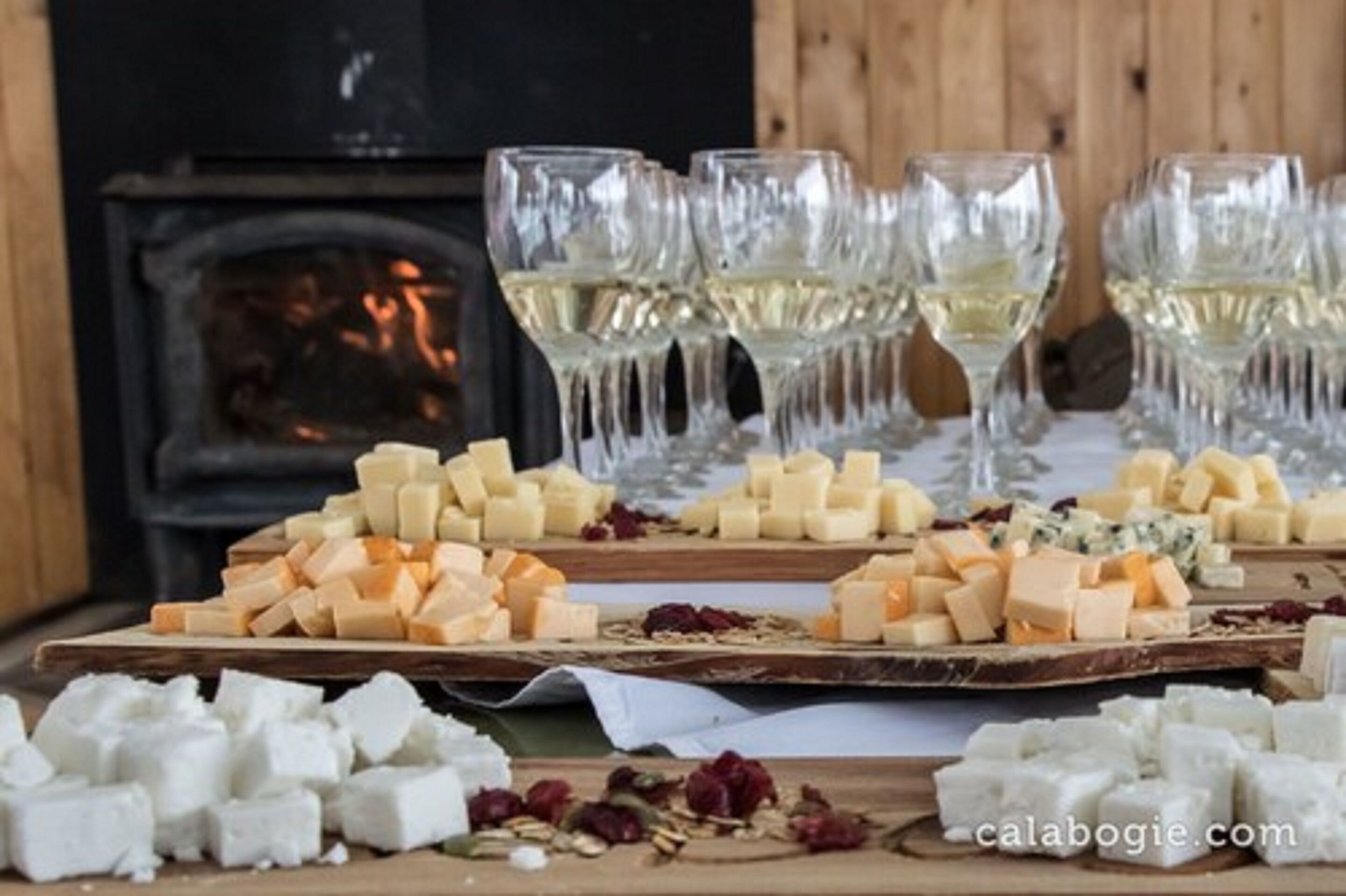 Eine Käseplatte mit verschiedenen Käsesorten und Trockenfrüchten, begleitet von Gläsern Weißwein, vor einem Holzhintergrund mit Kamin.