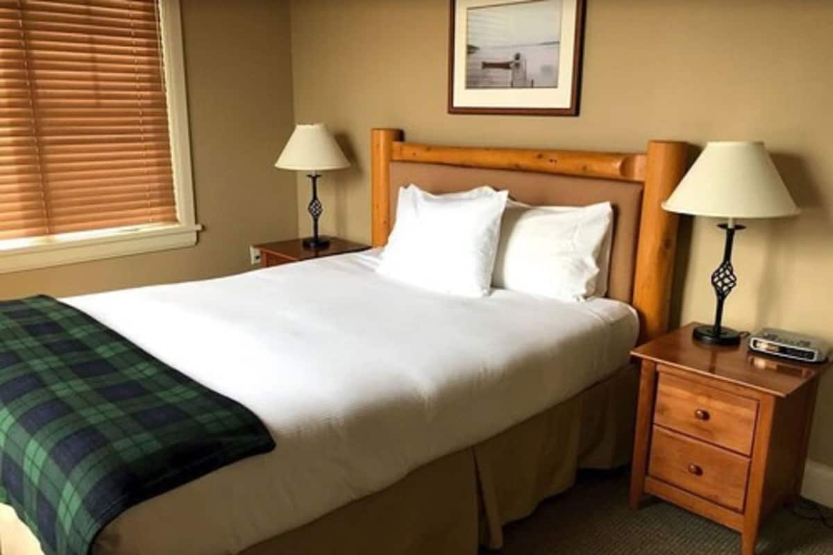 A hotel room with a large bed, two bedside tables with lamps, a window with blinds, and a framed picture on the wall.