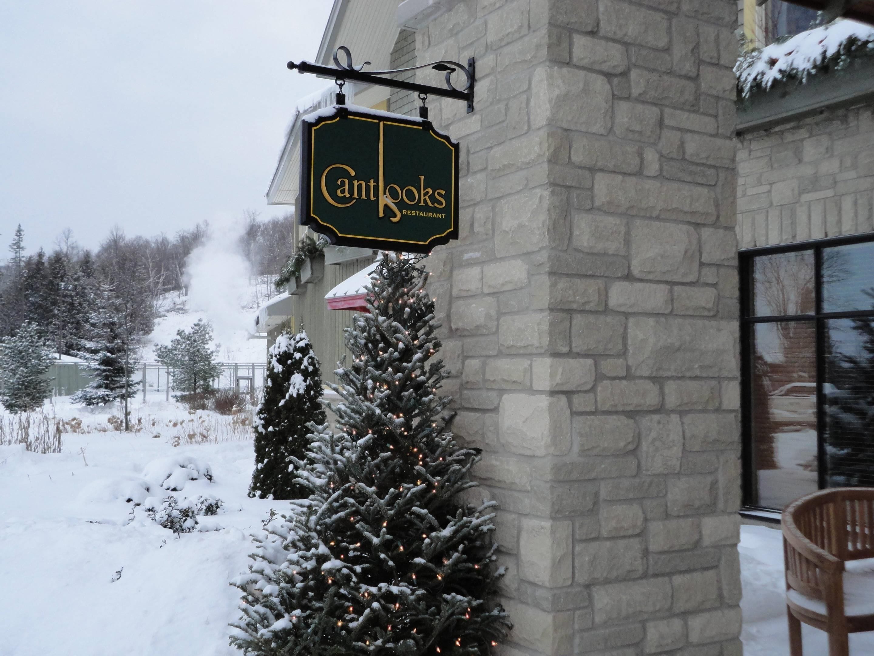 A sign for Cantooks Restaurant, a stone building with a snow-covered Christmas tree and a chair outside.