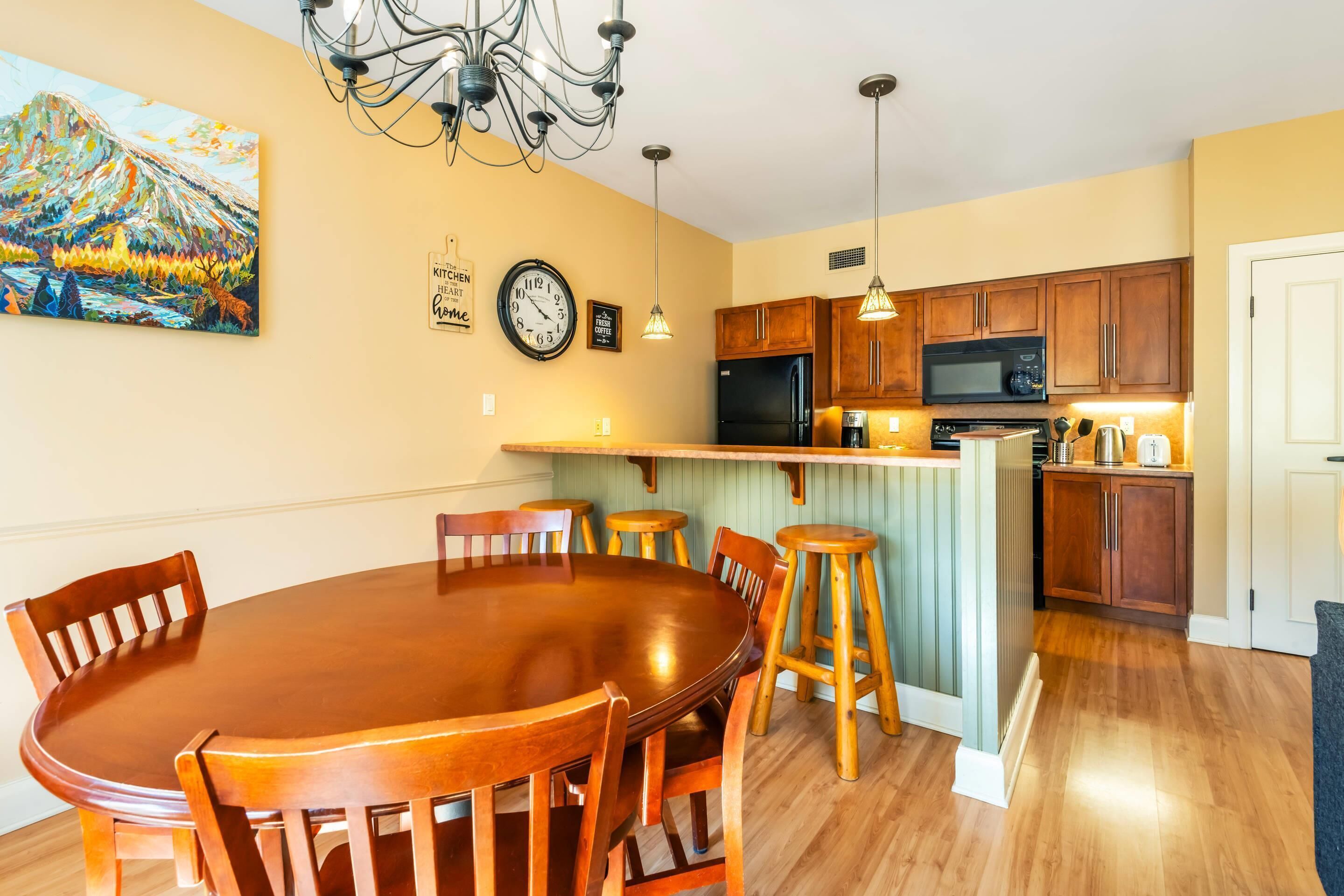 Un comedor con mesa y sillas de madera, una cocina con armarios y electrodomésticos de madera, un reloj y un cuadro en la pared.