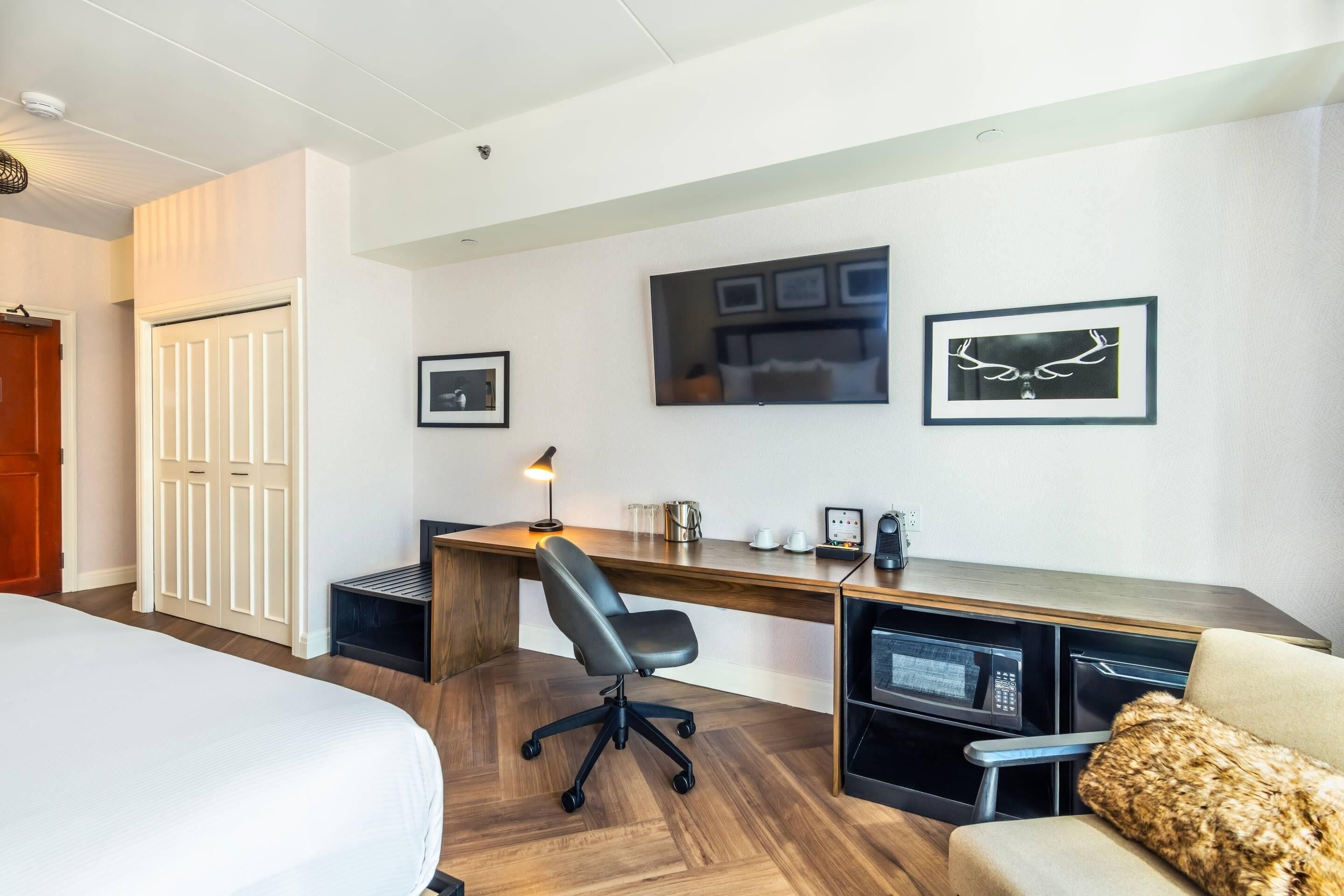 A hotel room with a bed, a desk with a chair, a microwave, a coffee maker, and framed pictures on the wall.