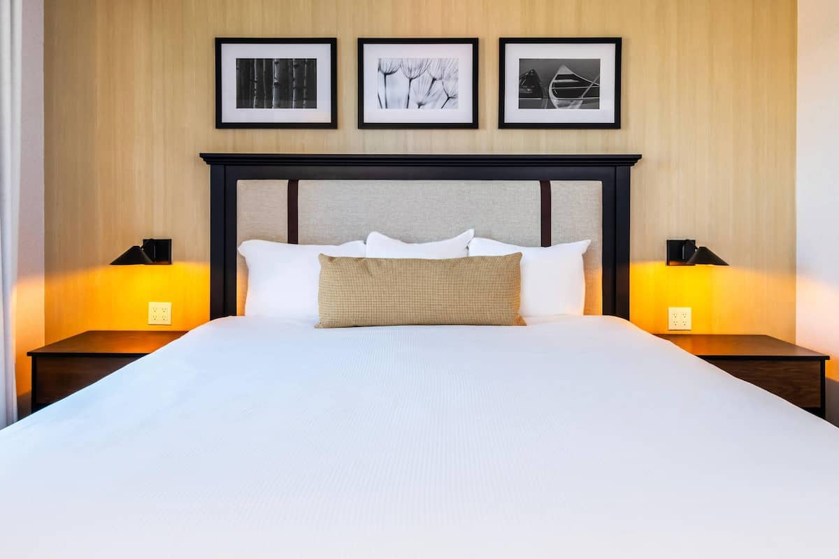 A neatly made bed with white linens and a checkered pillow, flanked by two bedside tables with lamps, against a wall with three framed pictures.