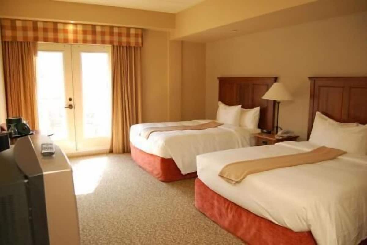 A hotel room with two beds, a desk, a lamp, and a view of the outdoors.