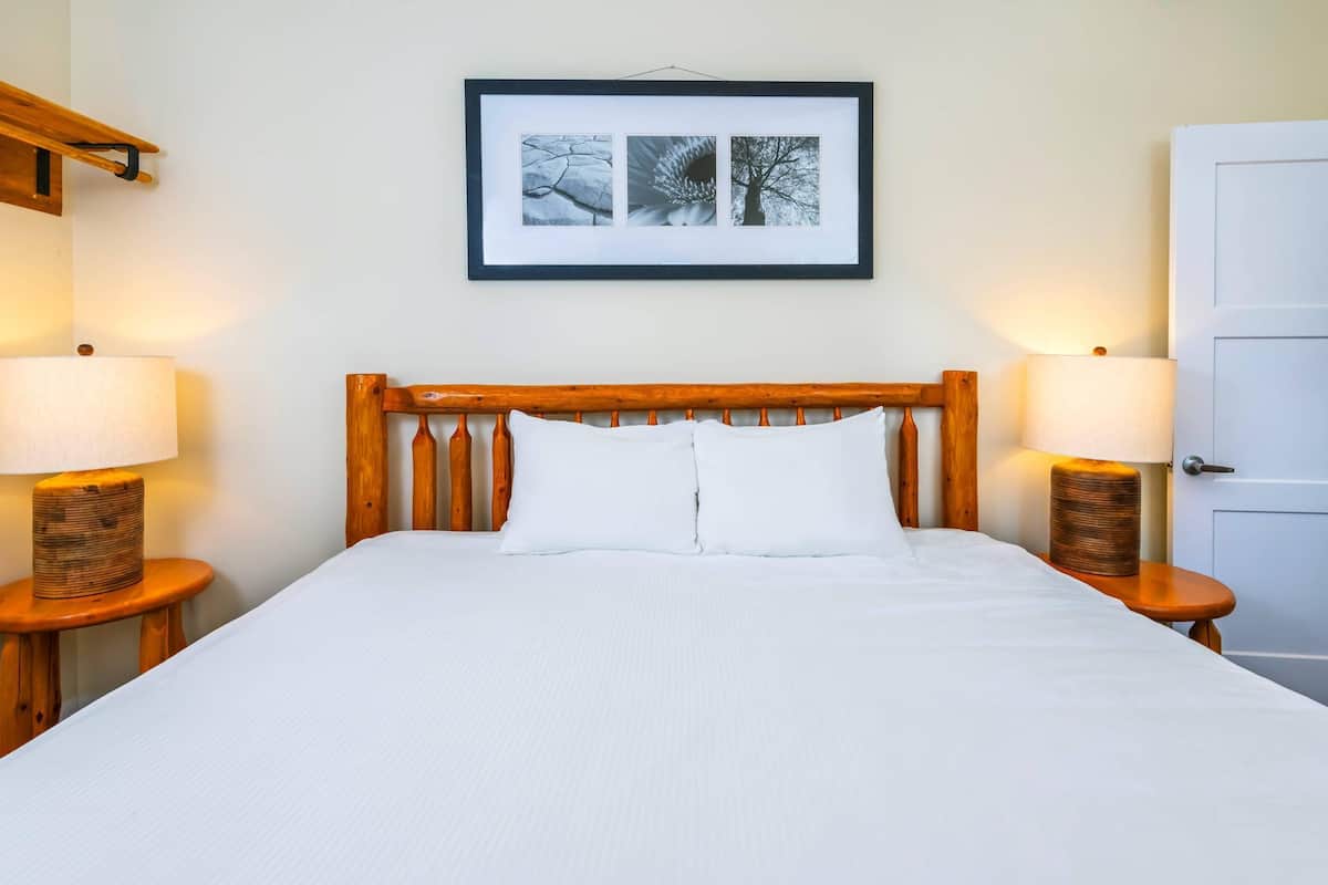 A neatly made bed with white linens, two bedside lamps, a framed picture above the bed, and a wooden headboard.