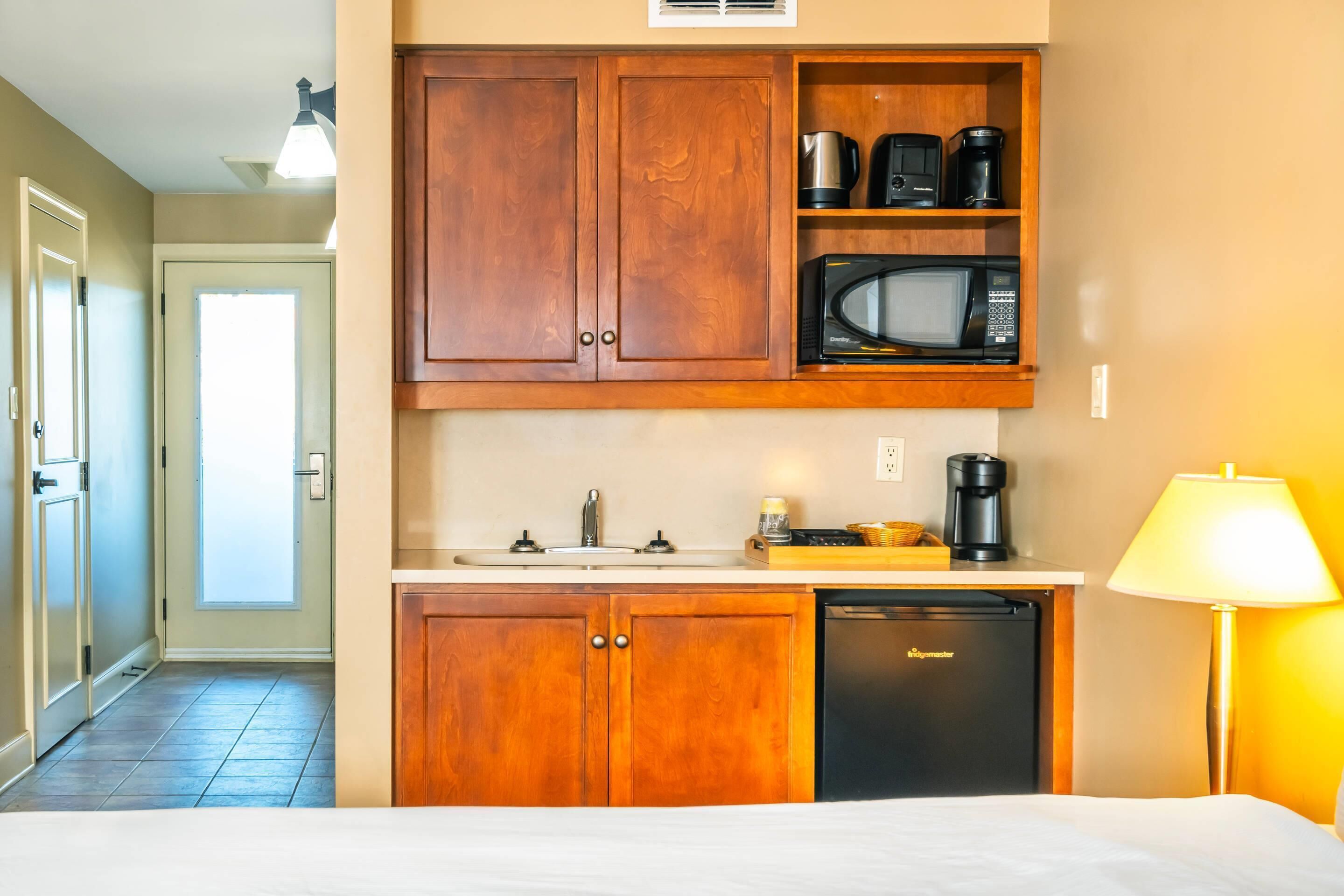 Une chambre d’hôtel compacte comprenant un coin cuisine, un lit, une lampe et une porte donnant sur un couloir.