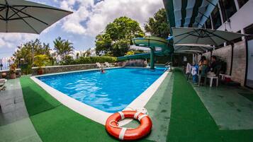 Uma piscina com escorrega, rodeada por guarda-sóis e espreguiçadeiras.