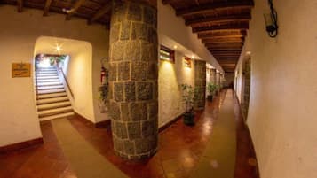 Un pasillo con pilares de piedra, vigas de madera y una escalera que lleva a un área exterior.