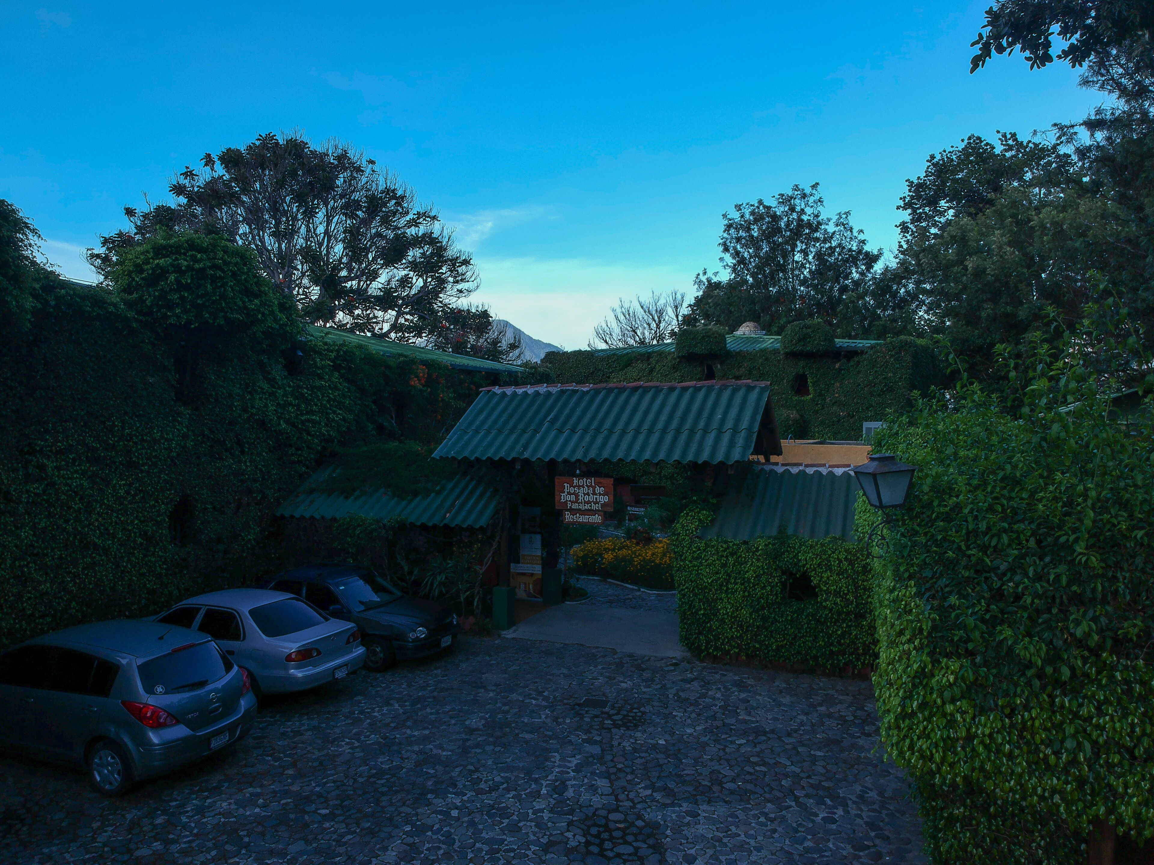 A rustic building with a green roof, surrounded by lush greenery. There are several parked cars in front.