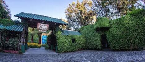 Un edificio rústico con techo verde, rodeado de abundante vegetación y un sendero adoquinado.