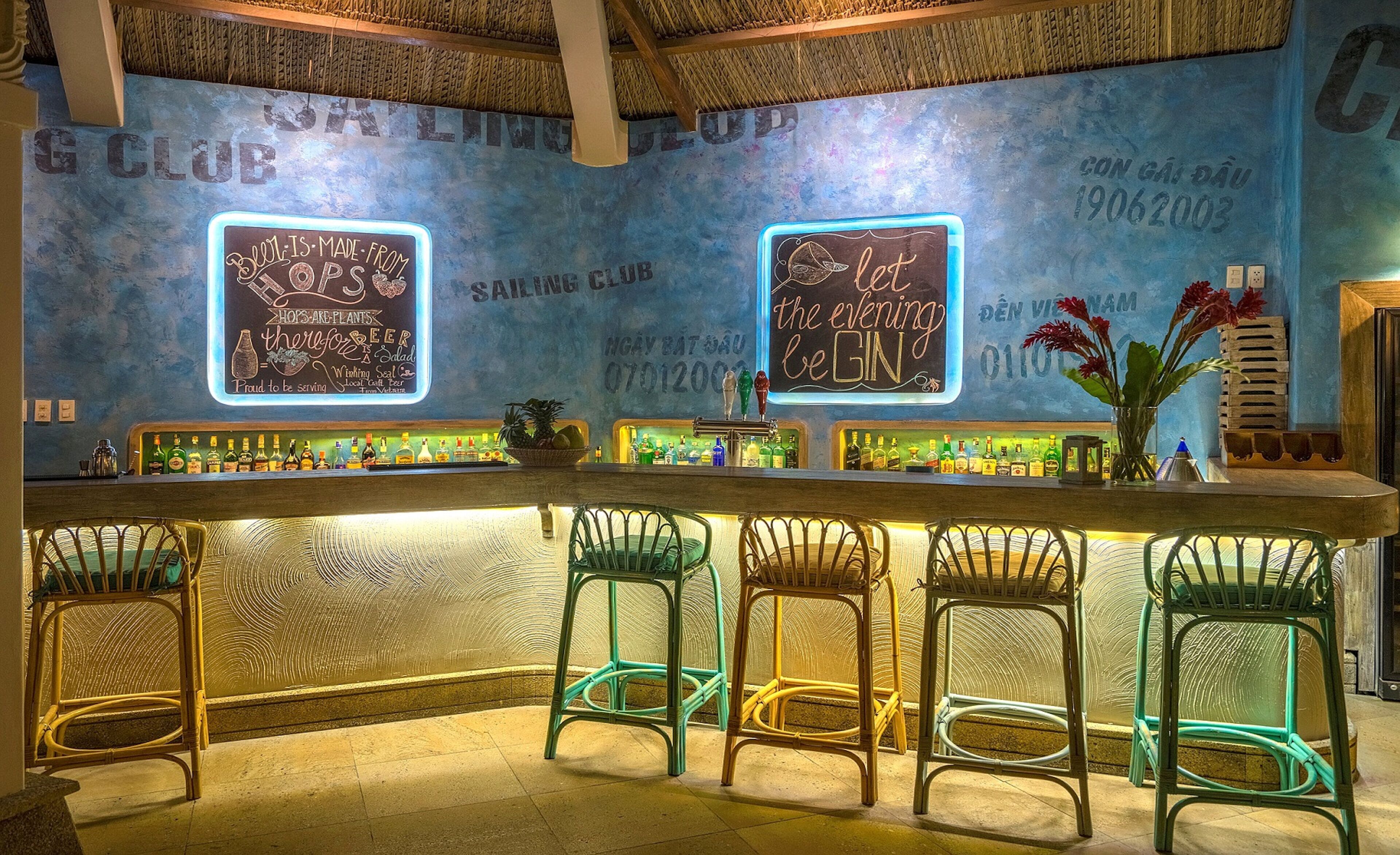 A bar area with neon signs, a variety of bottles, and colorful chairs.