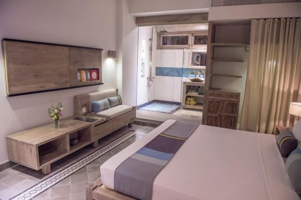 A modern hotel room with a large bed, a seating area with a sofa and a small table, a built-in TV, and a shower area.