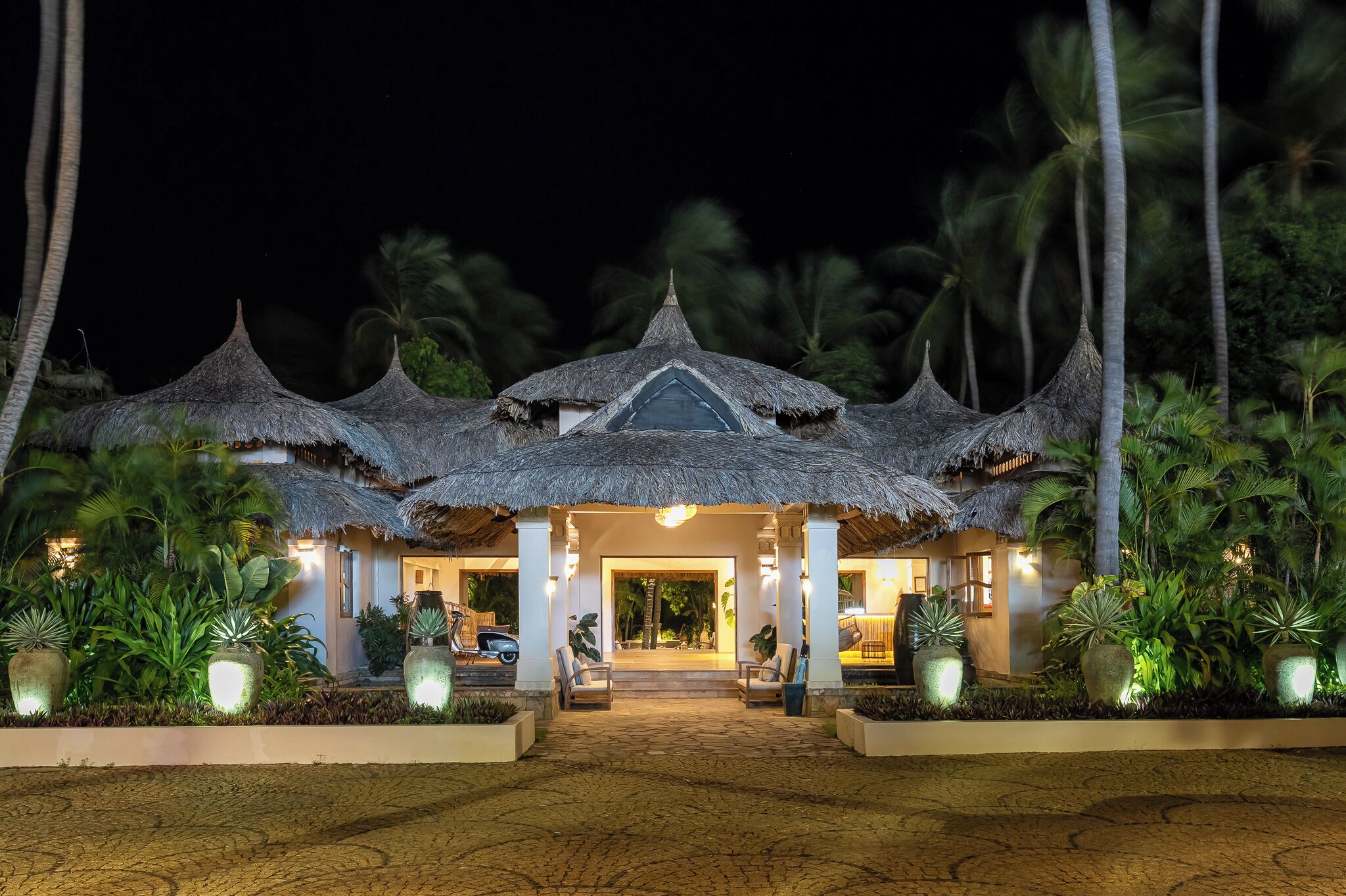 Un resort con edificios de techo de paja, entrada pavimentada y plantas tropicales.