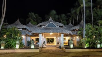 Un resort con edificios de techo de paja, entrada pavimentada y plantas tropicales.