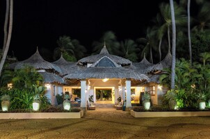 Un resort con edificios de techo de paja, entrada pavimentada y plantas tropicales.