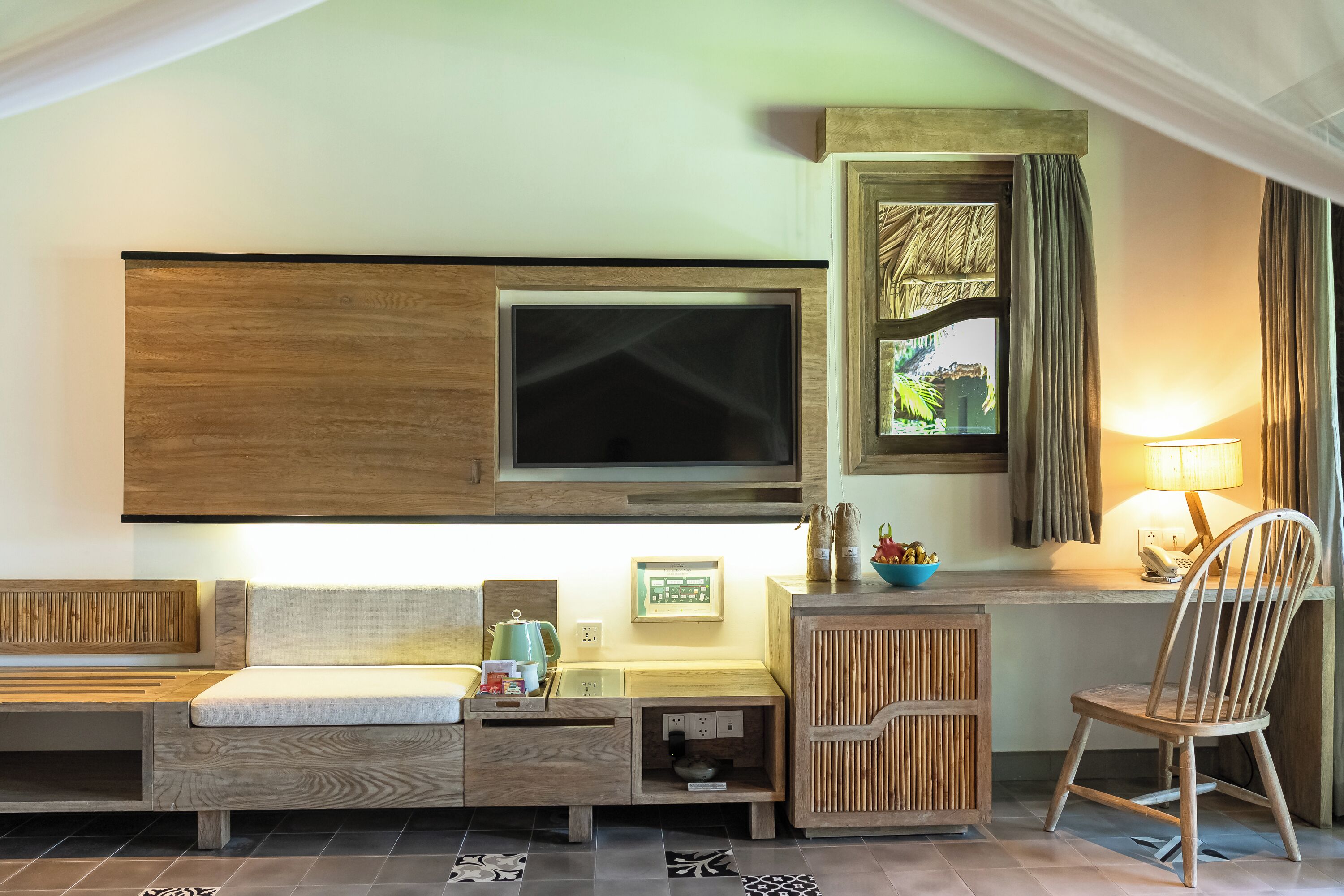 A hotel room with a TV mounted on the wall, a wooden desk with a chair, a small table with a lamp, and a window with curtains.
