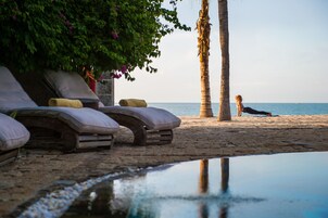Un espacio de relajación frente a la playa con sillas de descanso, una piscina pequeña y palmeras.