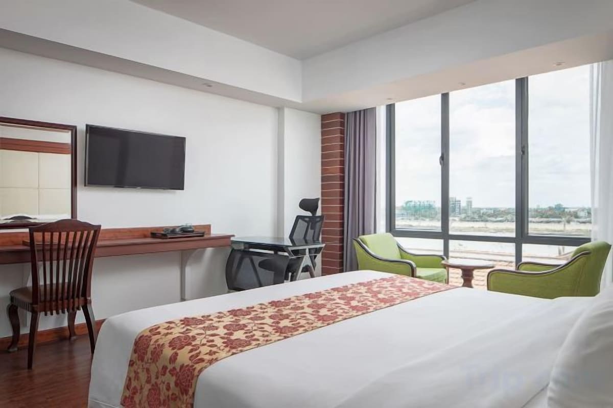 Une chambre d’hôtel équipée d’un lit, d’un bureau, de chaises, d’une télévision et d’une grande fenêtre donnant sur un paysage agréable.