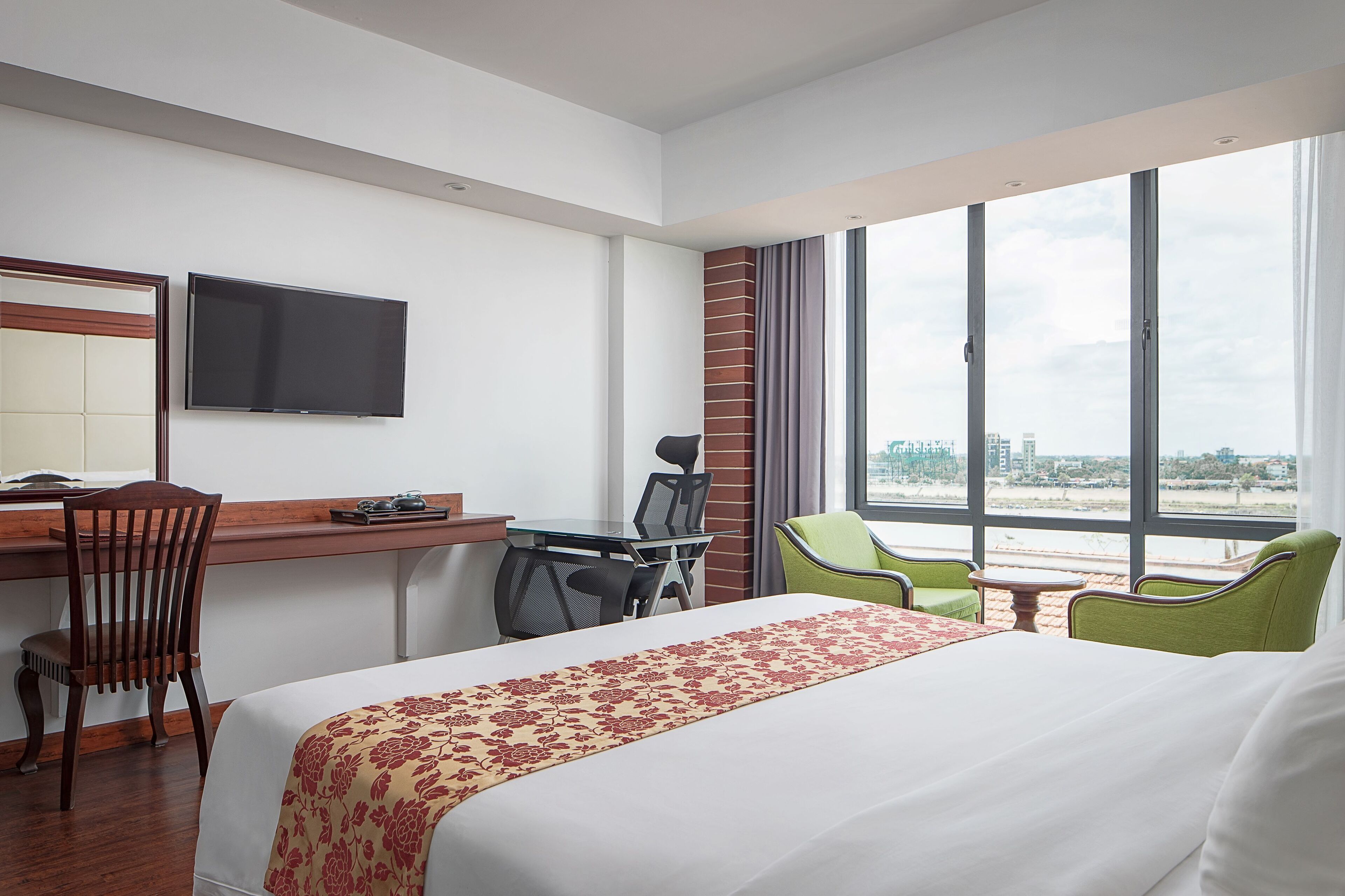A hotel room with a bed, desk, chairs, a TV, and a large window with a city view.