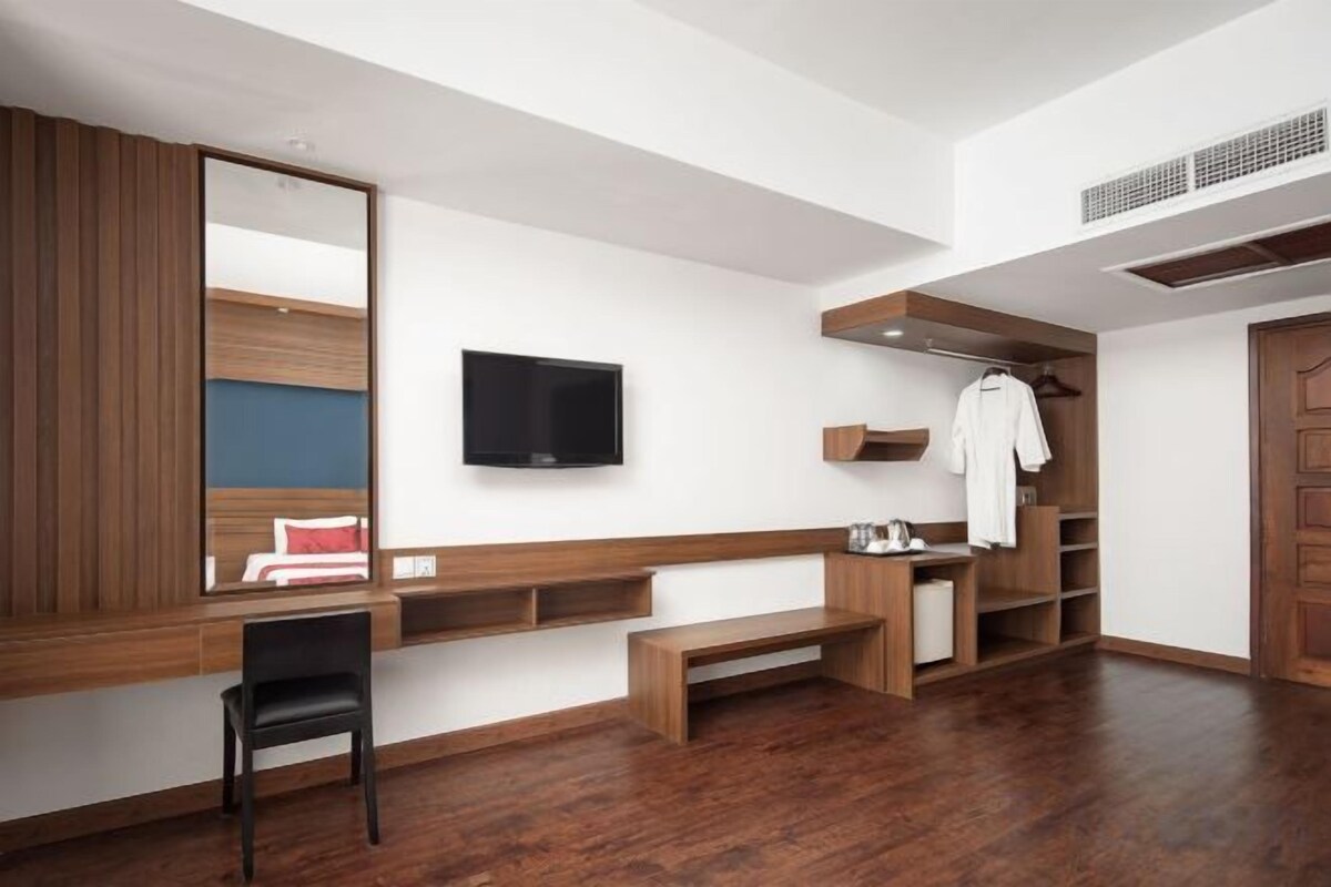 Habitación de hotel con un espejo grande, una televisión de pantalla plana, un banco de madera, un armario de madera y un banco con un cojín rojo.