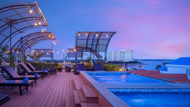 Un espace piscine aménagé sur le toit, avec des chaises longues et des parasols, offrant une vue sur un plan d’eau et le panorama de la ville.
