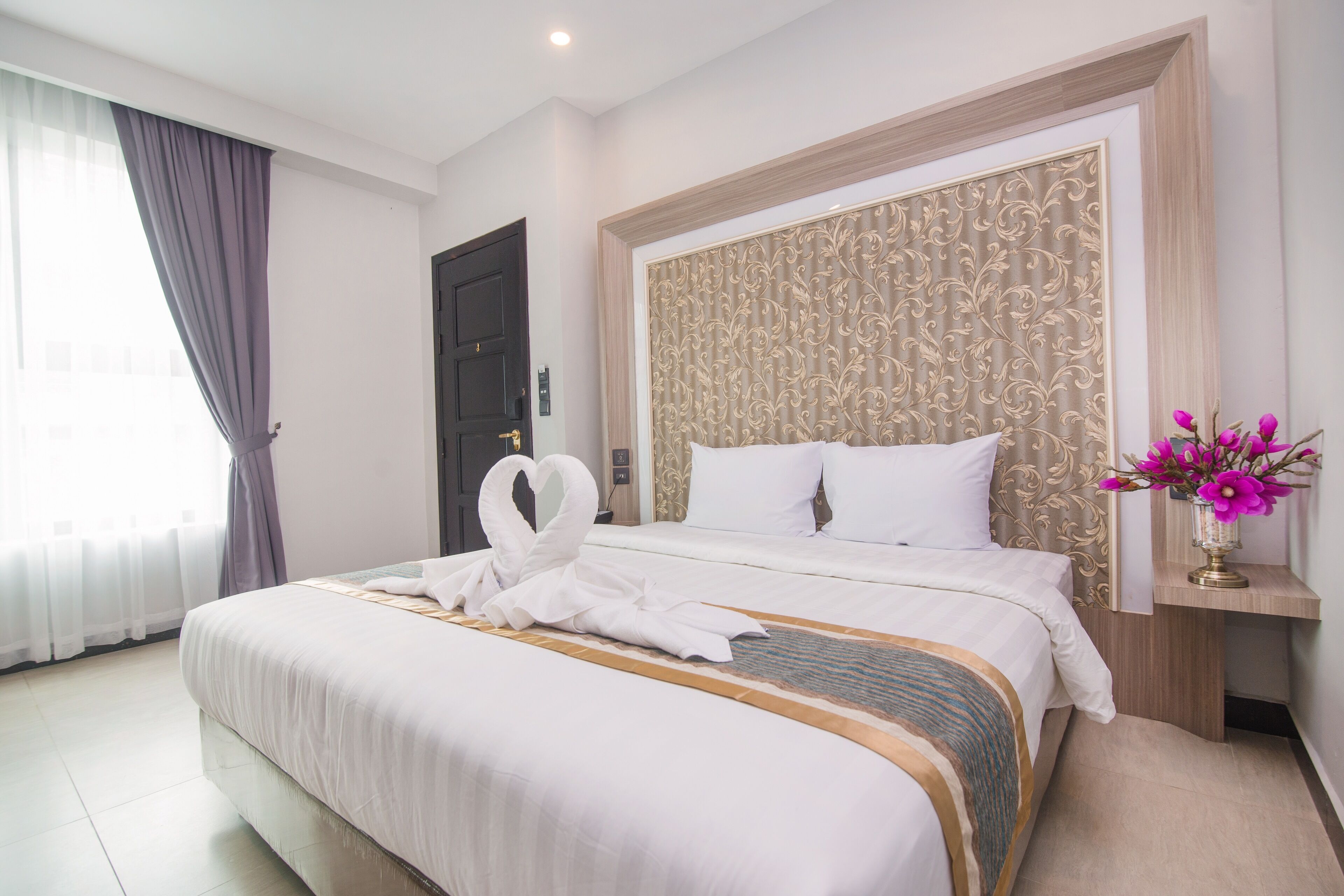 A hotel room with a large bed, a headboard with a decorative panel, a bedside table with a vase of flowers, and a window with curtains.