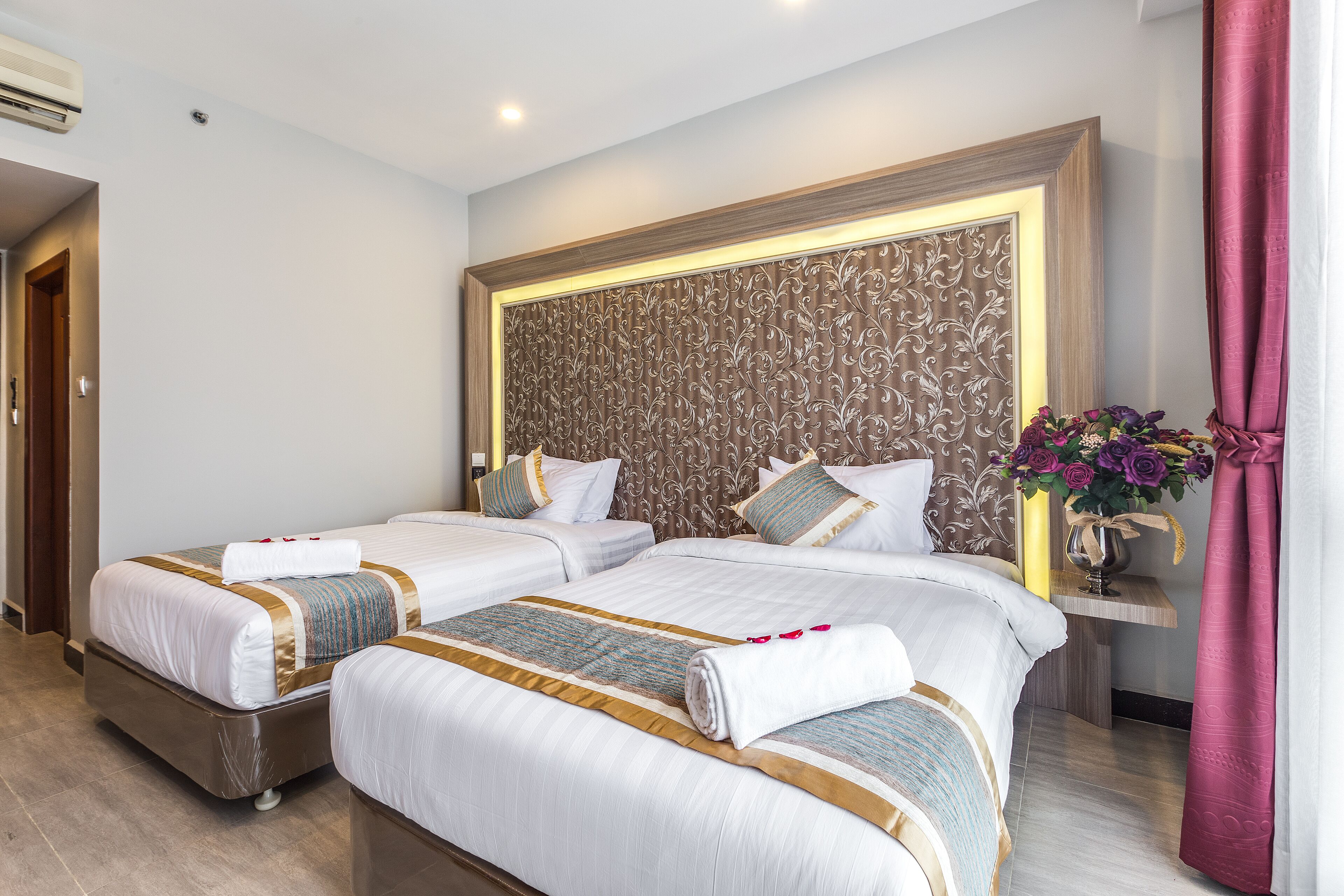 A hotel room with two beds, a wooden headboard, a nightstand with a vase of flowers, and a red curtain.