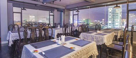 A restaurant with tables set for dining, featuring lace tablecloths and wooden chairs, with a cityscape view through large windows.