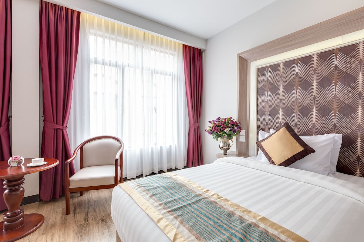 A hotel room with a large bed, a wooden chair, a small table, and a vase of flowers.