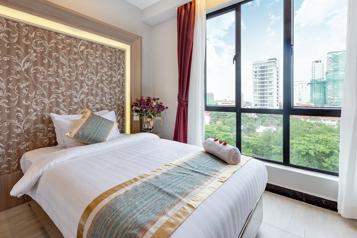 A hotel room with a large bed, a decorative headboard, a window with a city view, and a vase with flowers.