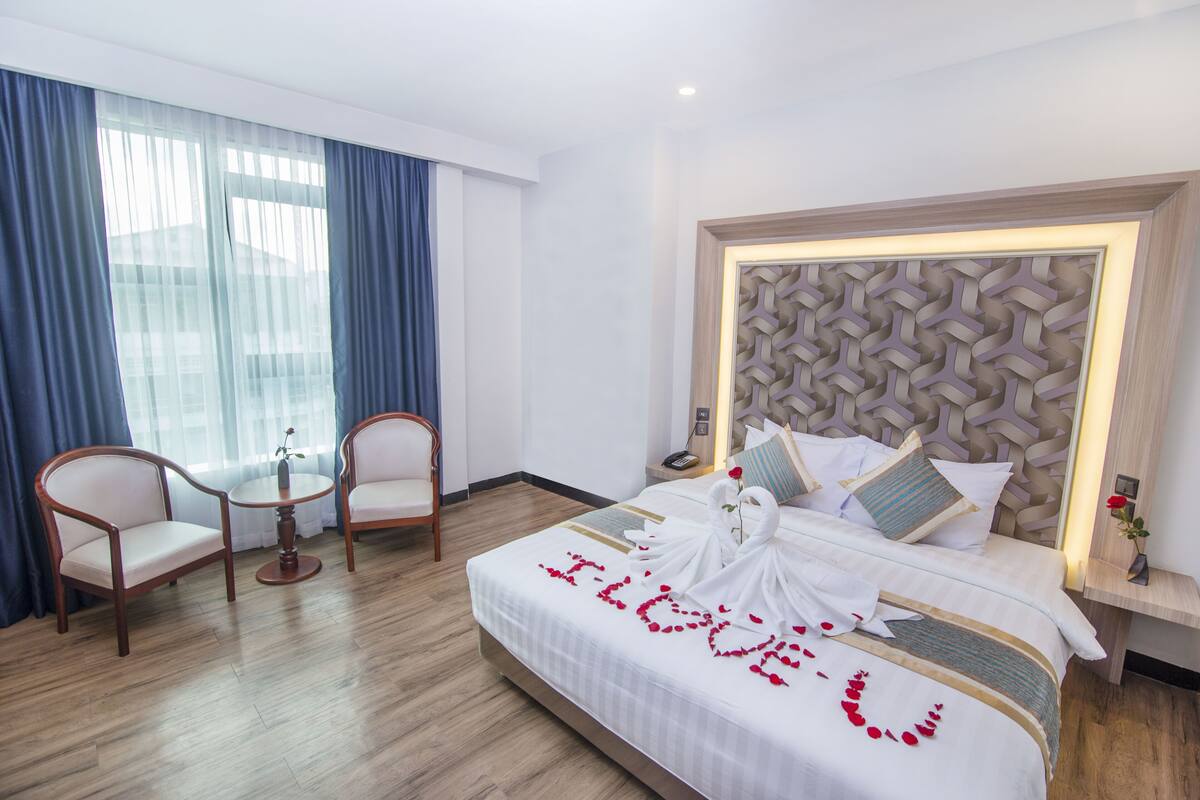A hotel room with a neatly made bed, two armchairs, a small table, and a decorative headboard.