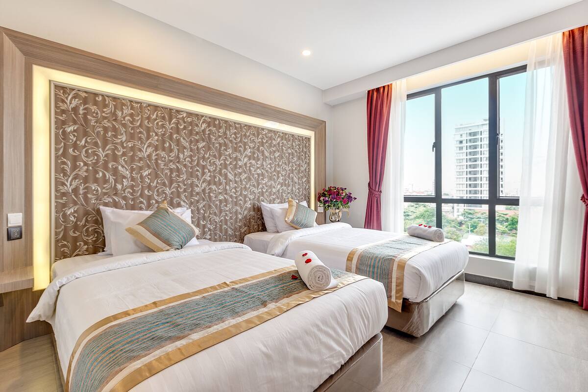 A hotel room with two beds, a decorative headboard, a large window with curtains, and a view of buildings.