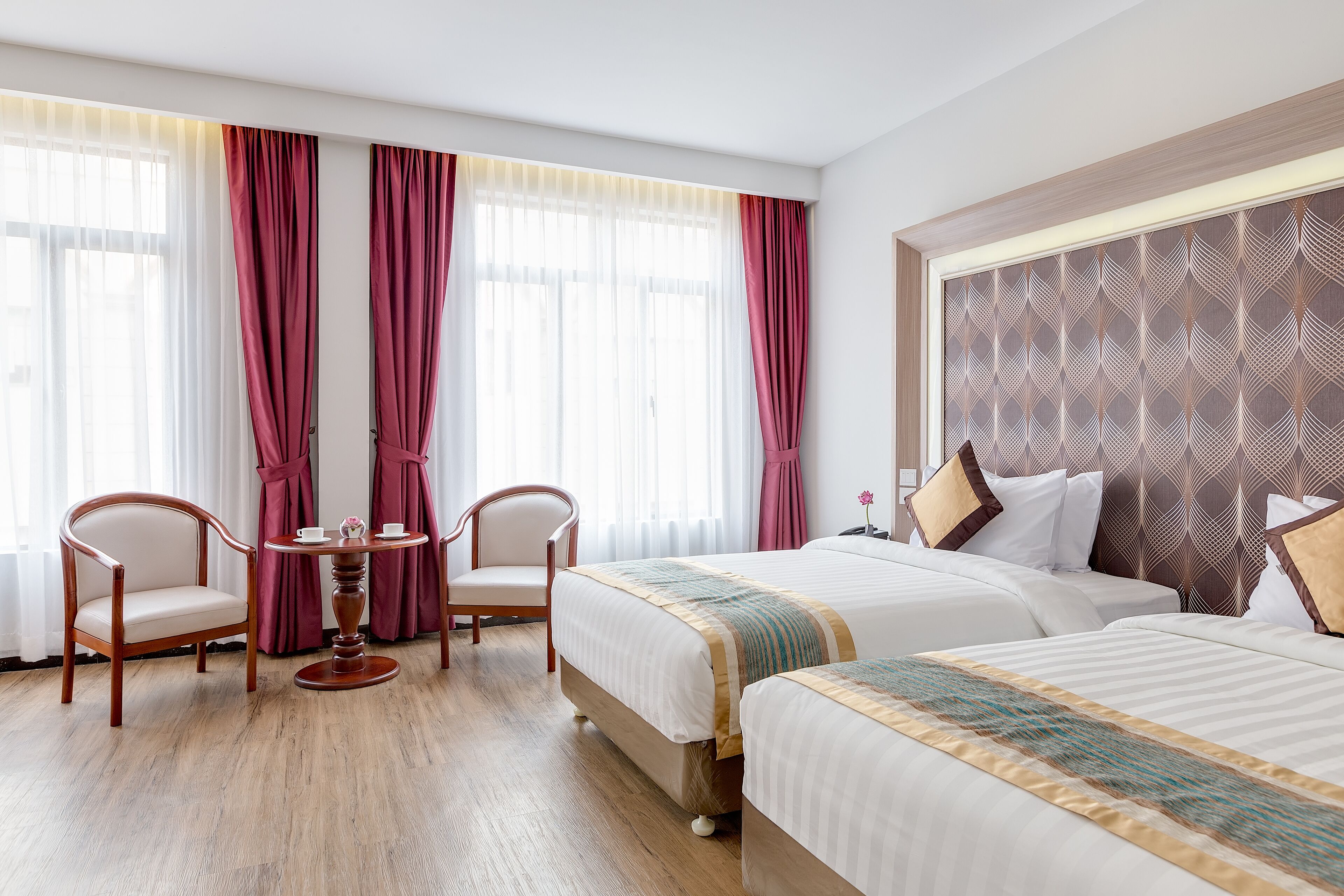 A hotel room with two beds, a wooden chair, a small round table, and a large window with curtains.