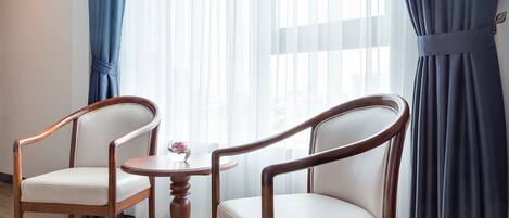 A room with two wooden chairs and a small round table, a window with sheer curtains, and a view of the city.