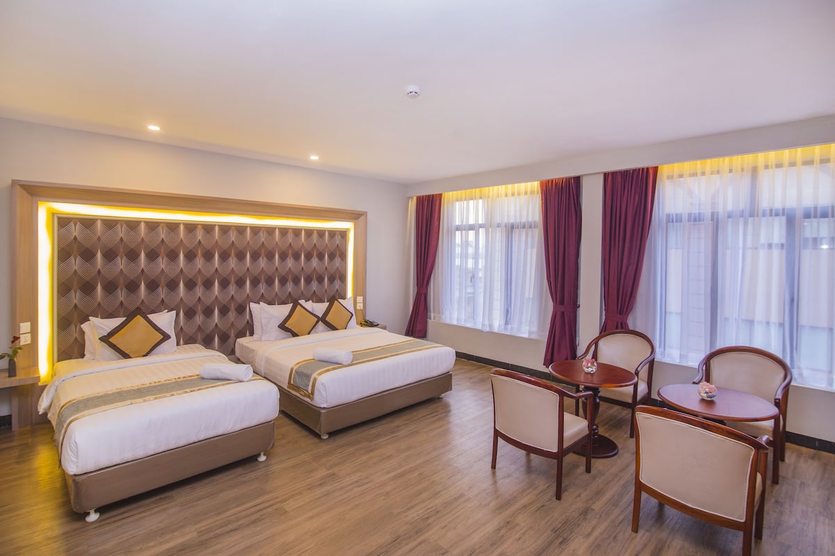 A hotel room with two beds, a sitting area with a table and chairs, and large windows with curtains.