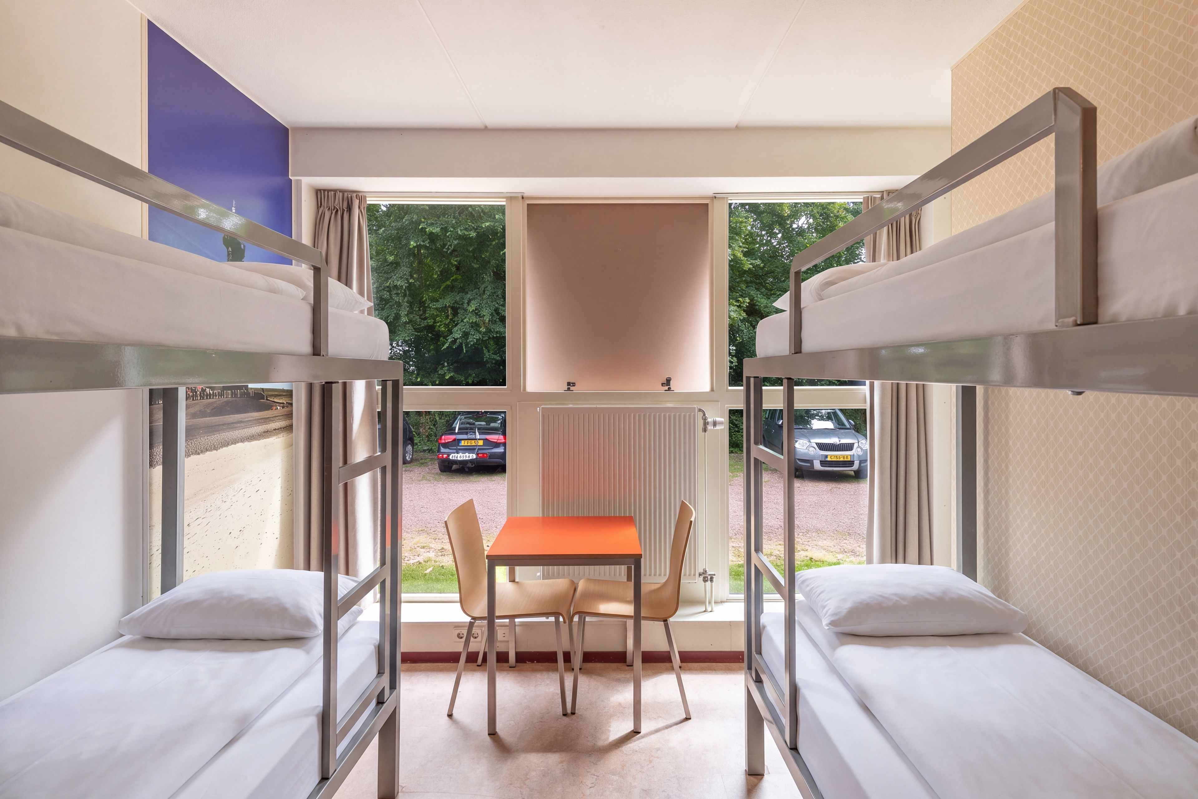 A dormitory room with bunk beds, a table, and chairs, and a view of parked cars outside.