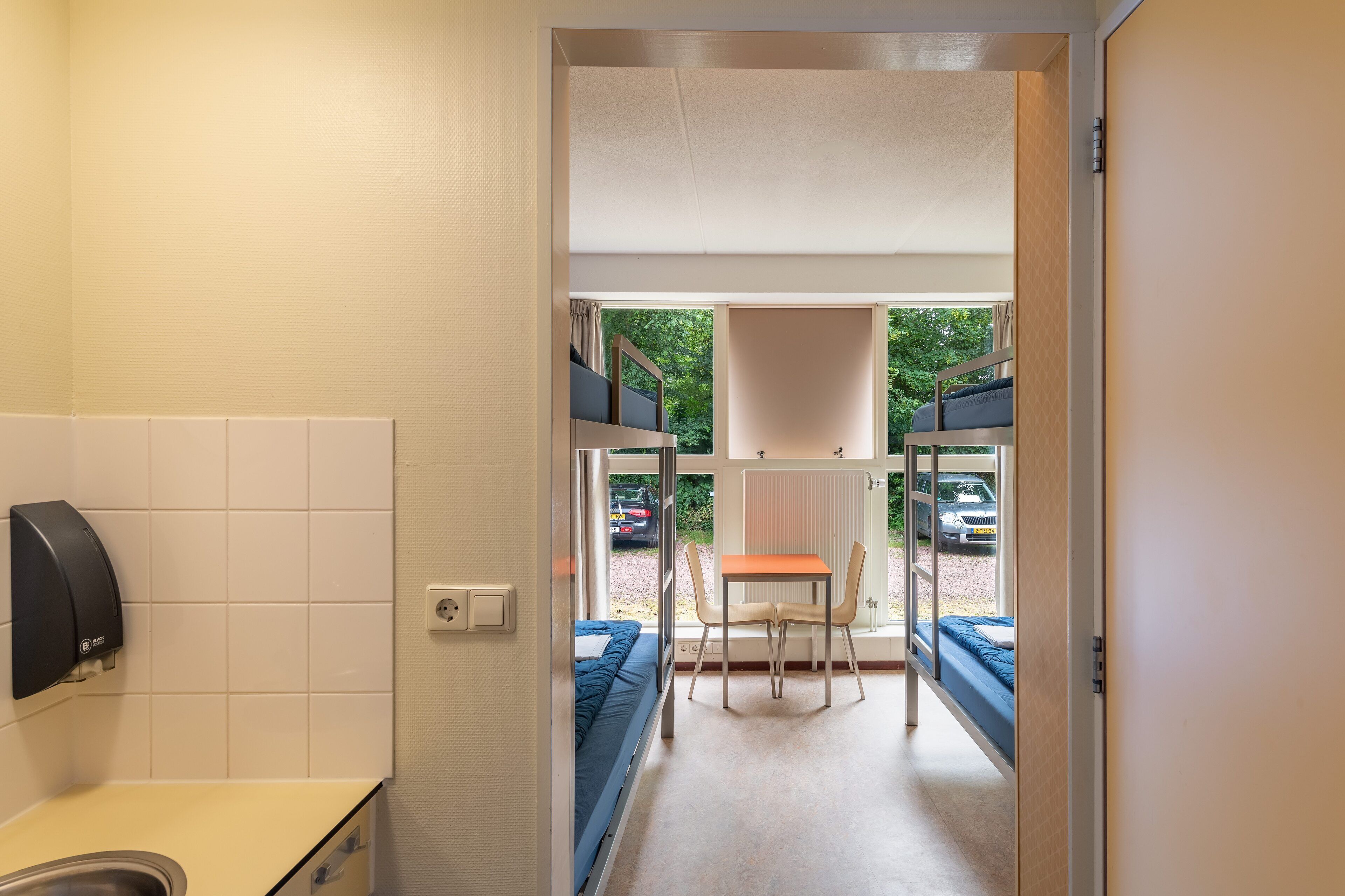 Une chambre d’hôtel avec un lit, une table et des chaises, une fenêtre donnant sur les voitures garées et la verdure à l’extérieur.