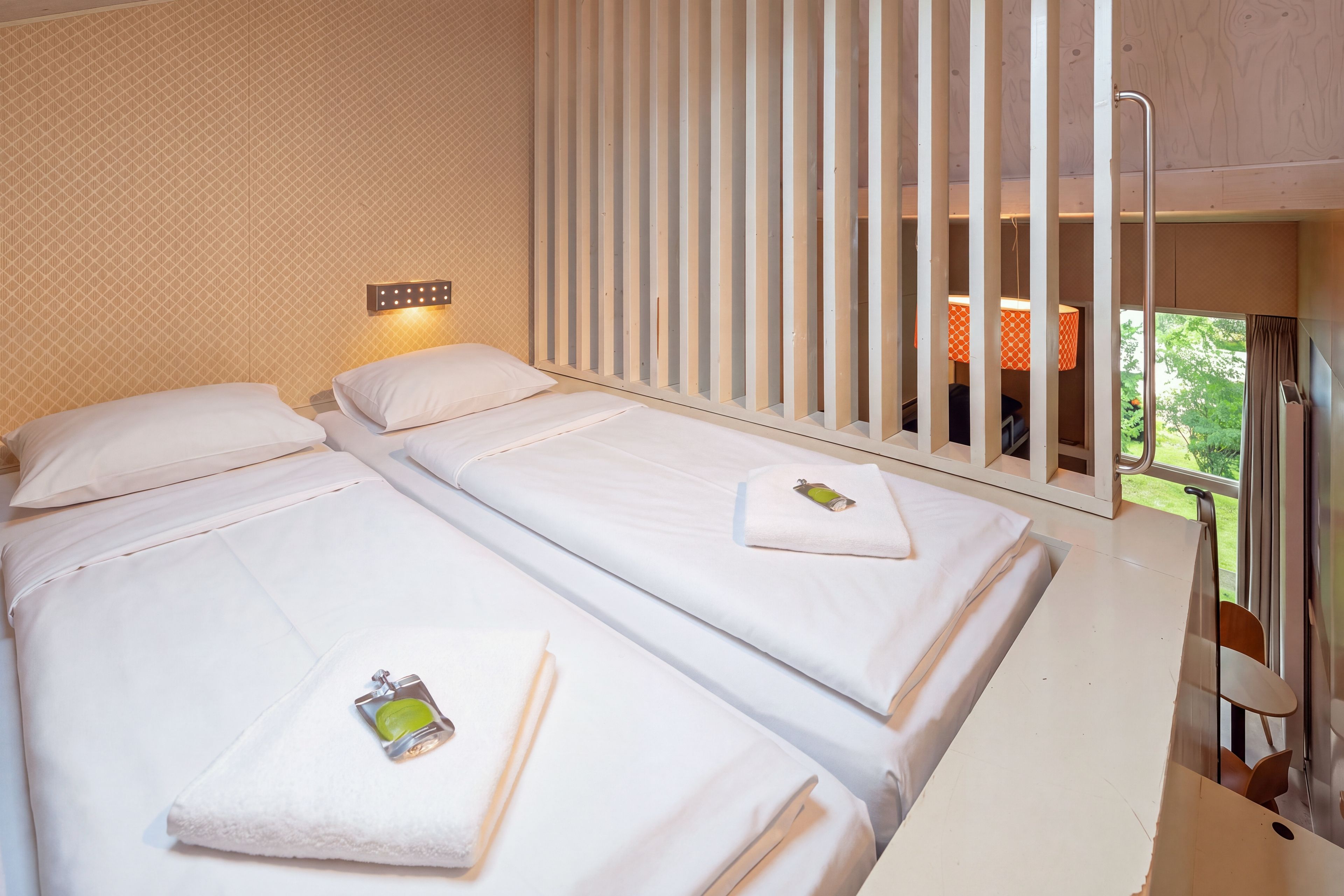 A hotel room with two beds, a window with curtains, and a view of greenery outside.