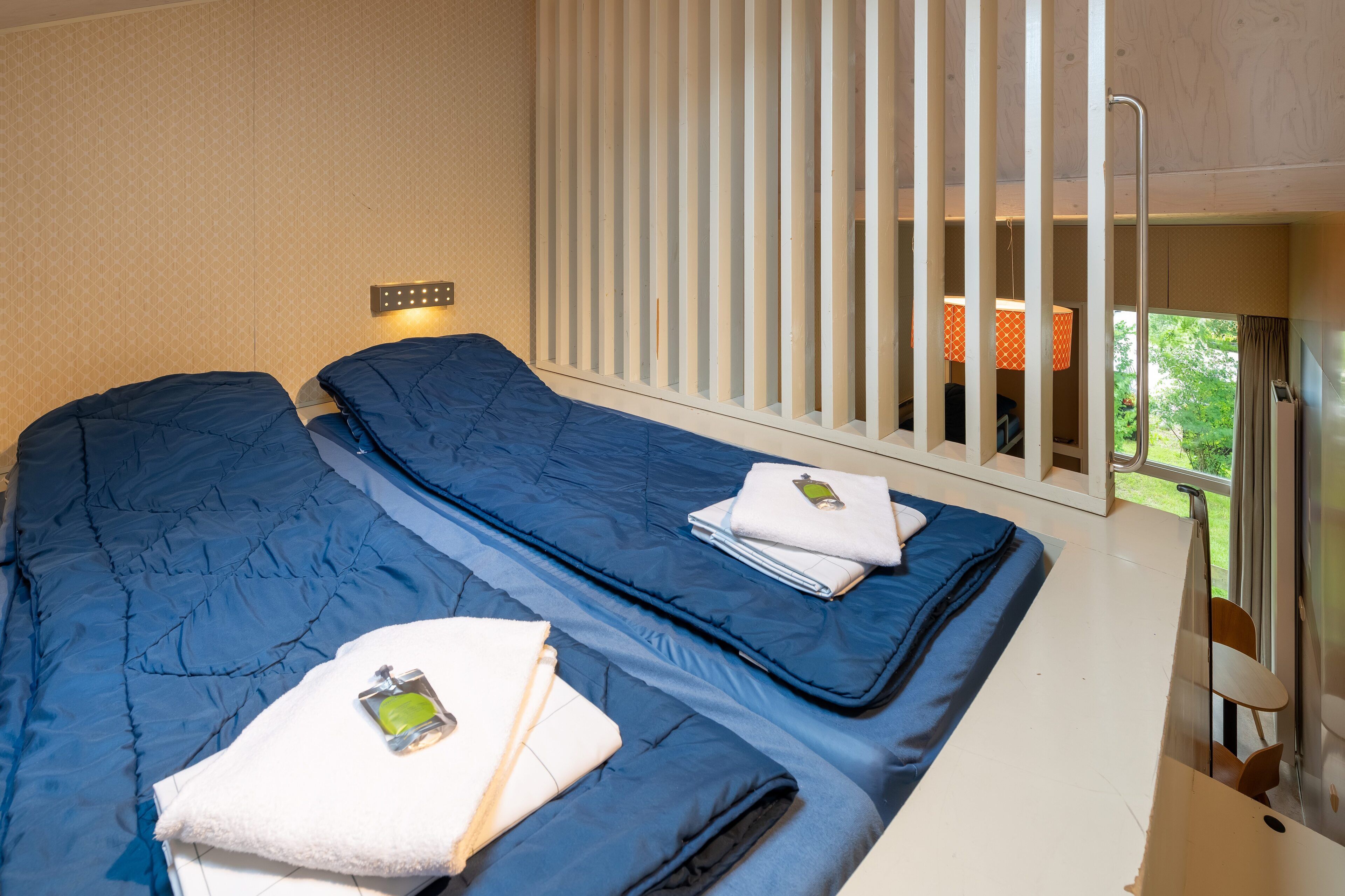 A hotel room with two beds, a window with blinds, a small table, and a view of greenery outside.